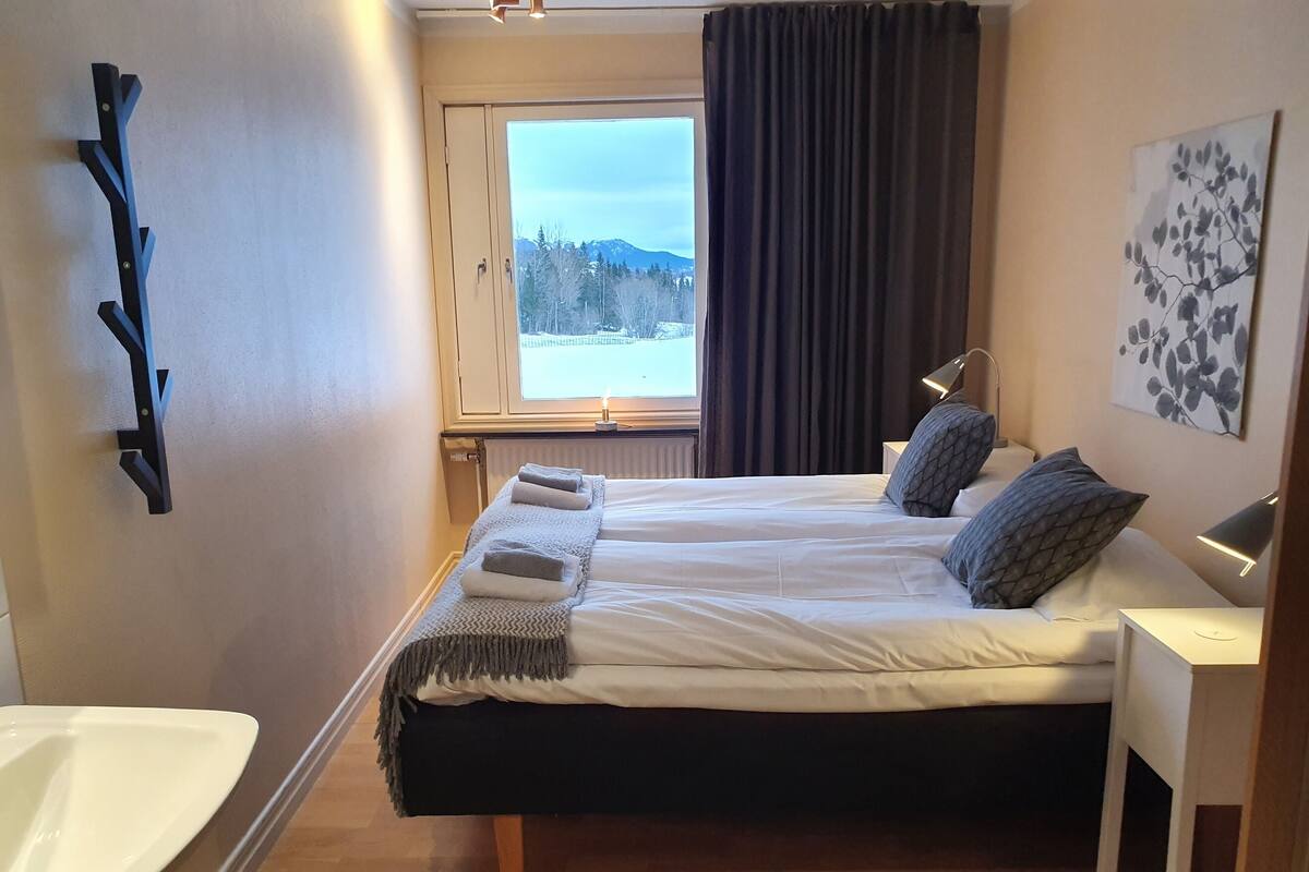 Un dormitorio con cama, mesitas de noche, ventana con vistas, estante en la pared y un cuadro.