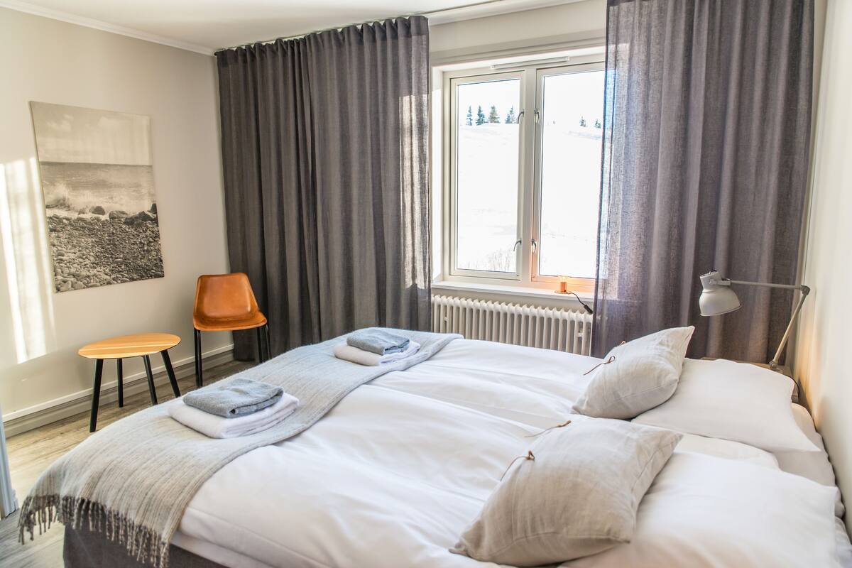 Una habitación de hotel con cama, ventana con cortinas, radiador, una silla y un cuadro en la pared.