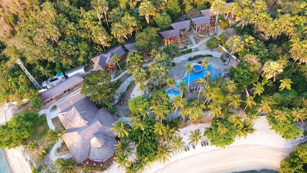 Vue aérienne d’un complexe hôtelier comprenant plusieurs bâtiments de style bungalow, une piscine centrale et des palmiers environnants.