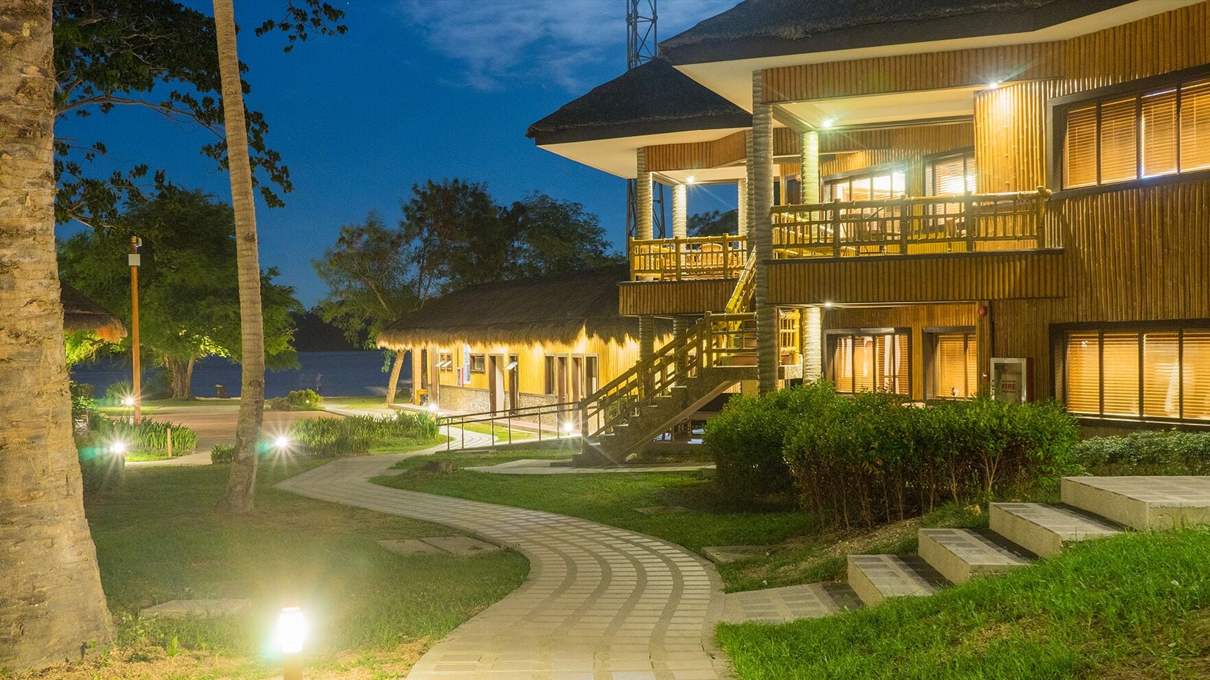 Un resort bien iluminado con un edificio de techo de paja, una escalera que lleva a un balcón y un camino pavimentado.