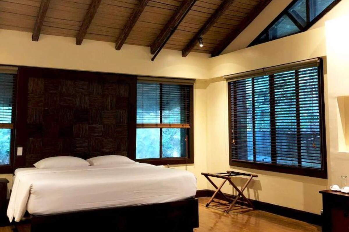 Un dormitorio con una cama grande, vigas de madera en el techo y amplias ventanas con persianas.