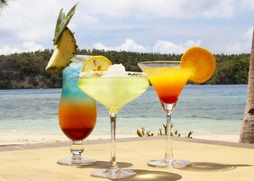 Tres cócteles tropicales sobre una mesa de madera, con vista a una playa y al océano.