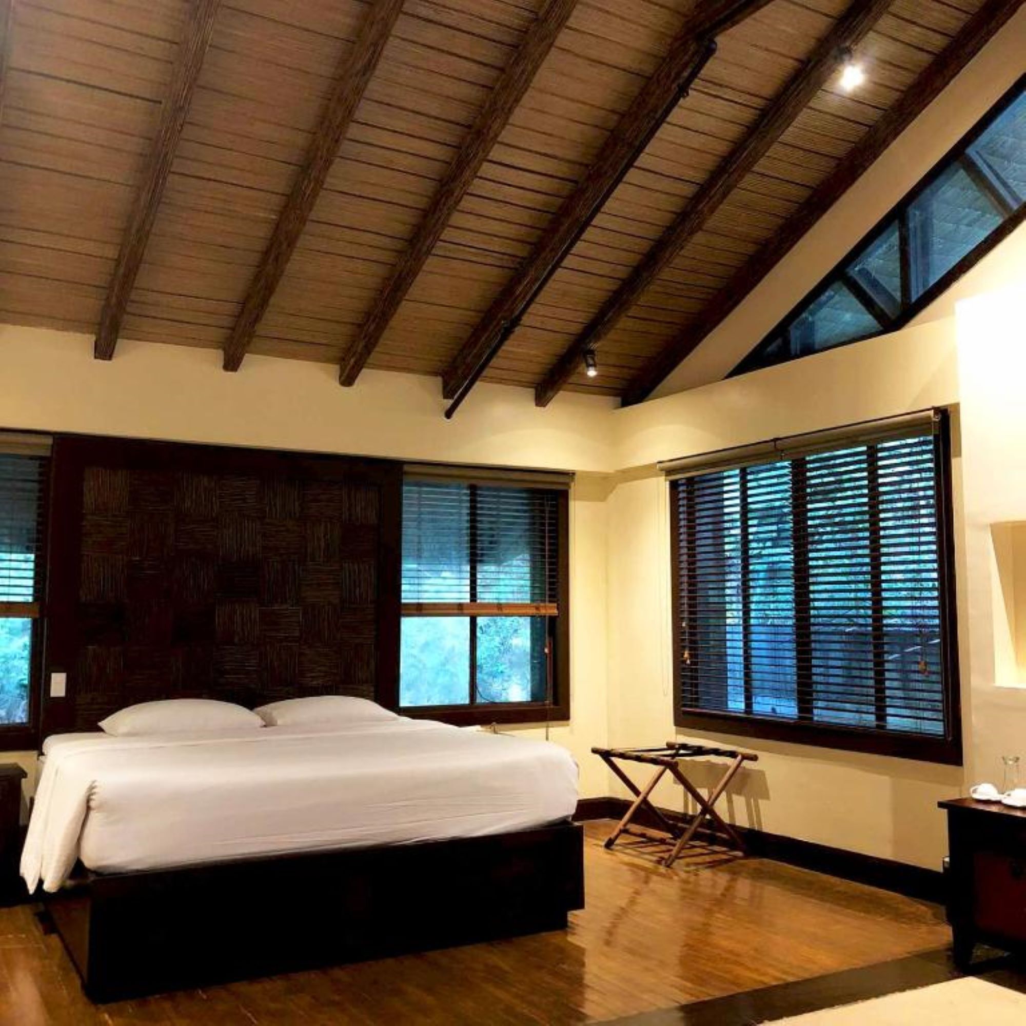 A bedroom with a large bed, wooden ceiling beams, and large windows with blinds.