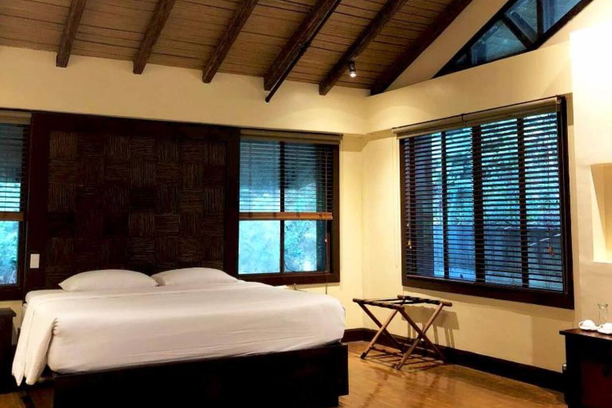 Une chambre avec un grand lit, des poutres apparentes en bois au plafond et de grandes fenêtres équipées de stores.