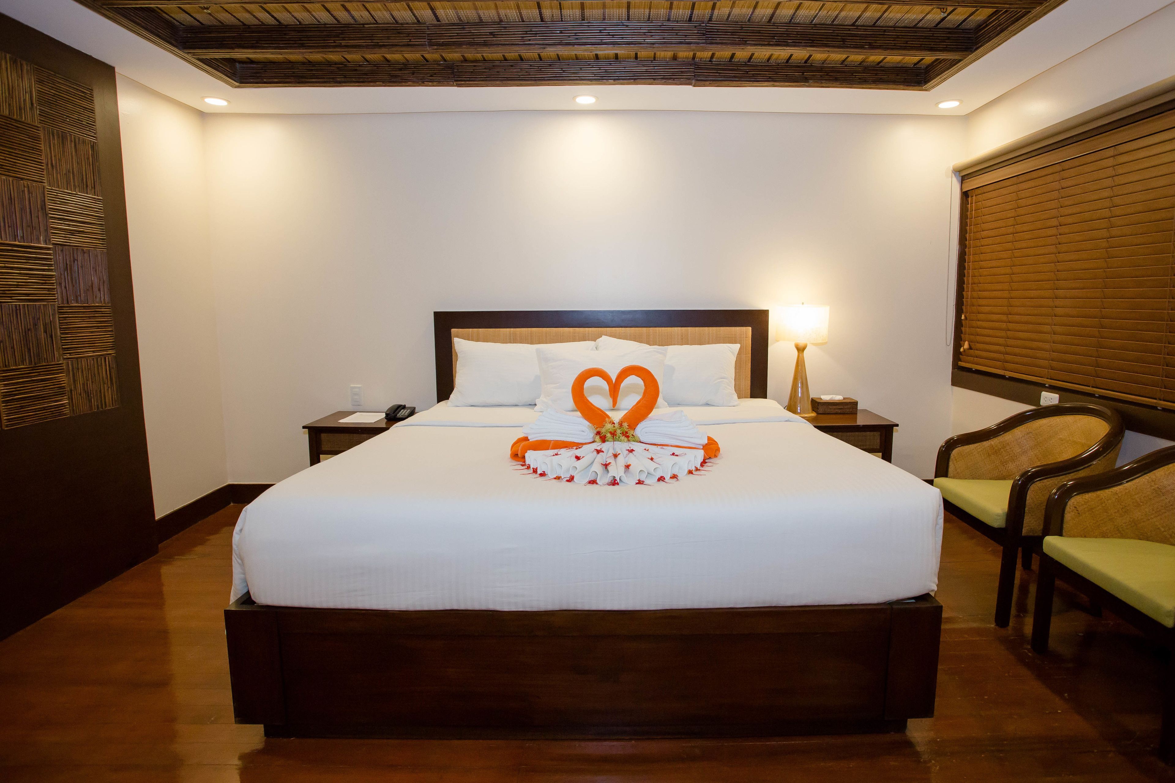 A neatly made bed with a heart-shaped towel arrangement, a bedside lamp, and a wooden headboard.