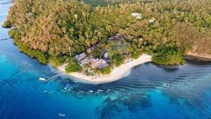 An aerial view of a tropical island with a white sandy beach, dense green forest, and several buildings with thatched roofs.