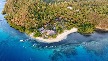 Vista aérea de una isla tropical con una playa de arena blanca, un bosque verde frondoso y varias edificaciones con techos de paja.