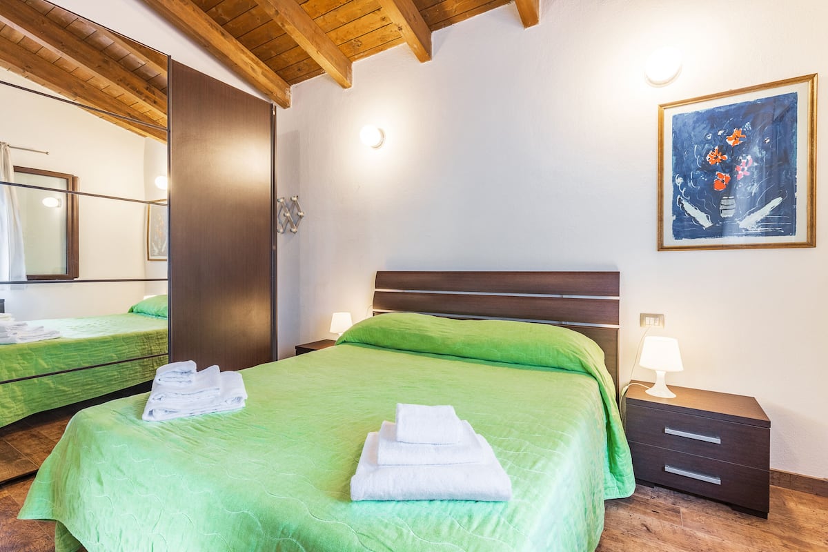 A hotel room with two beds, green bedding, a wooden ceiling, a painting on the wall, and a nightstand with a lamp.
