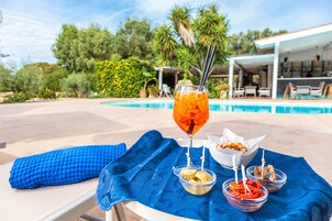 Escena junto a la piscina con una toalla azul, un vaso de bebida helada y dos recipientes pequeños de comida sobre una mesa.