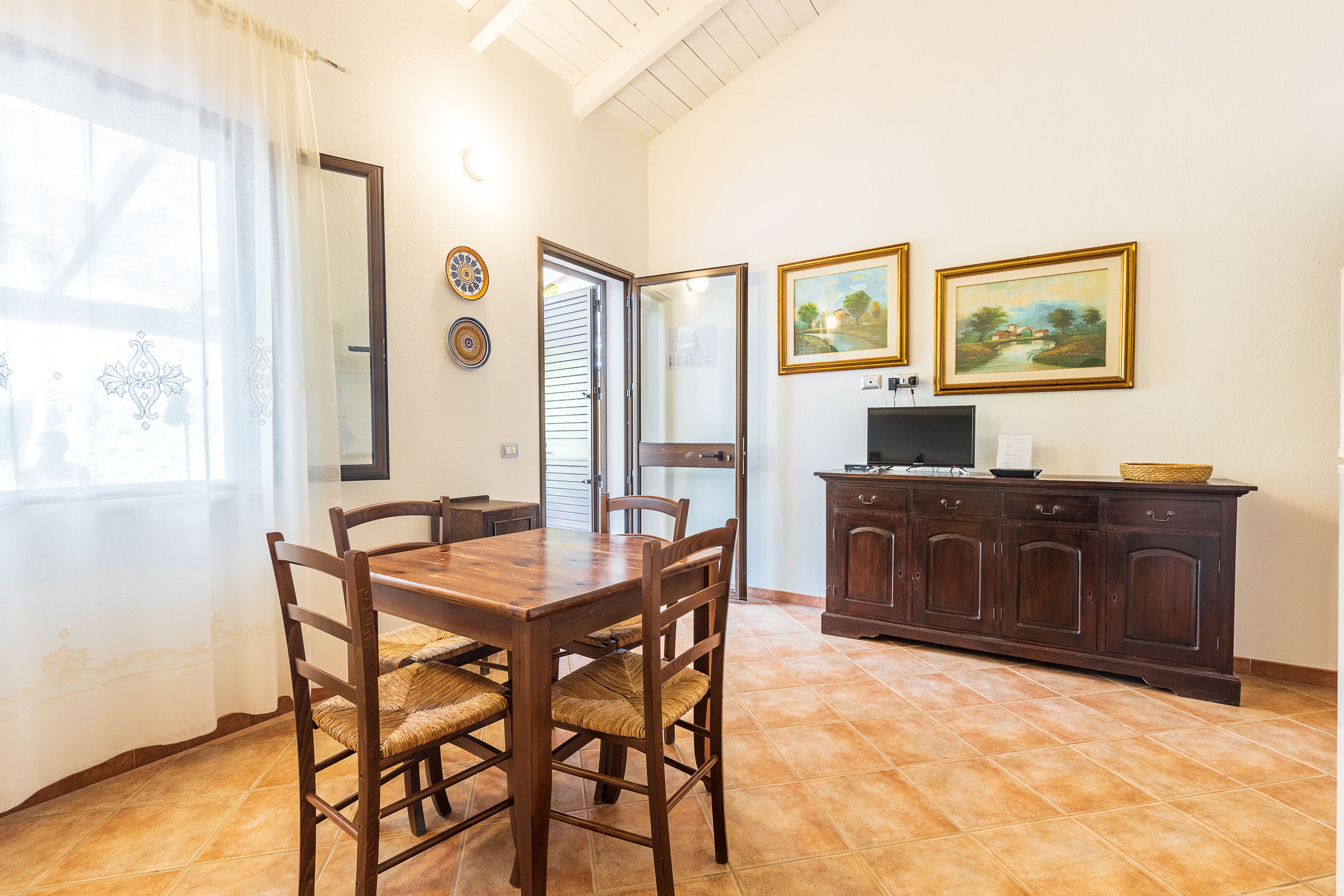A dining area with a wooden table and chairs, a cabinet with a television, and two framed paintings on the wall.