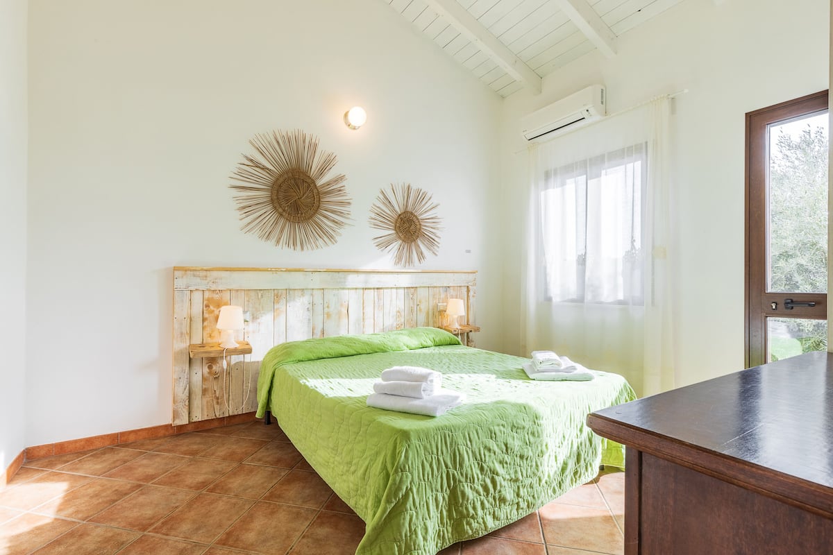 A bedroom with a wooden headboard, a green bedspread, two wall-mounted decorative sunburst mirrors, a bedside table with a lamp, and a view of greenery through a window.