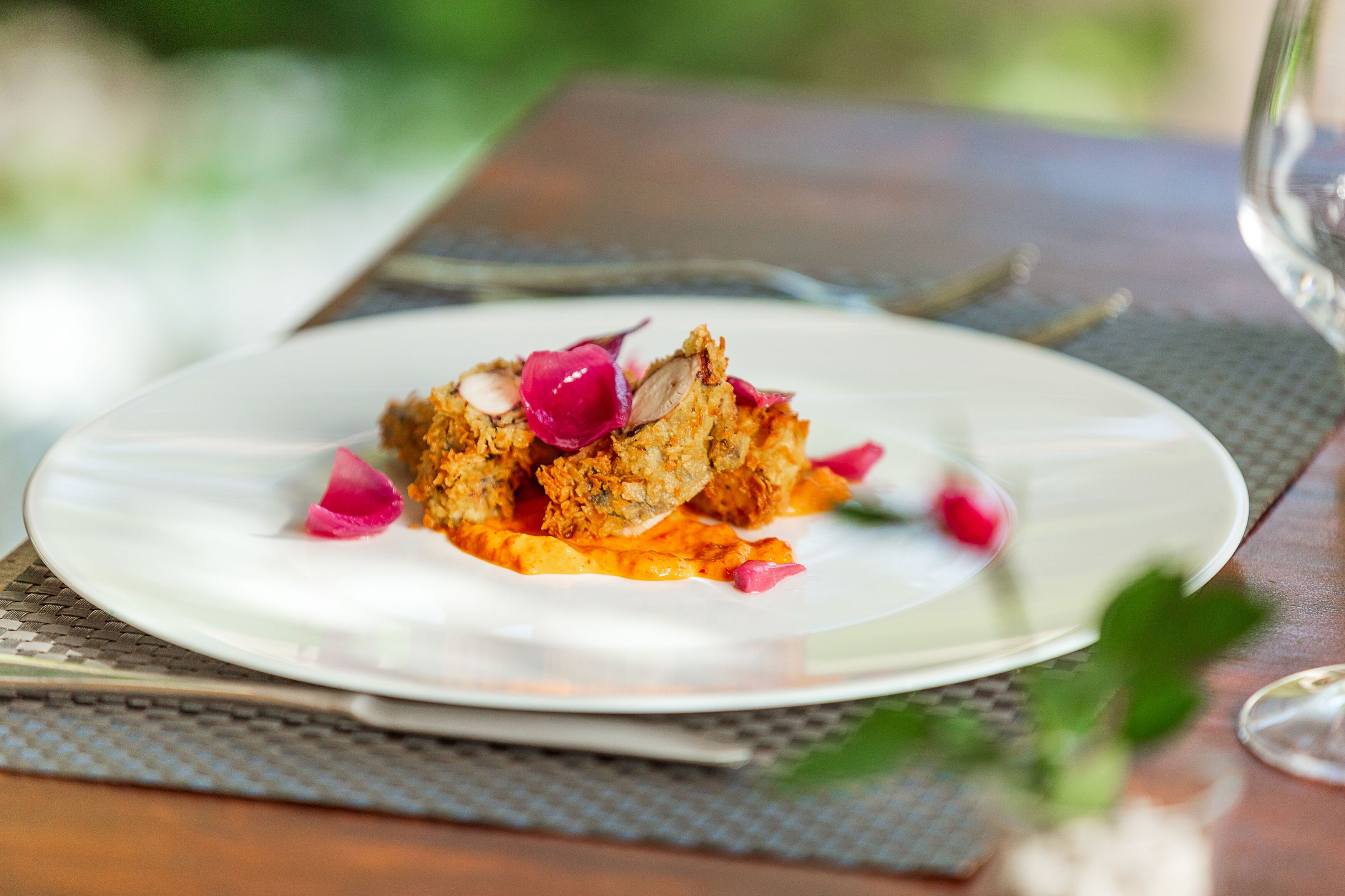 A plate of food with a white rim, garnished with pink petals and a small pink flower, on a textured table mat.