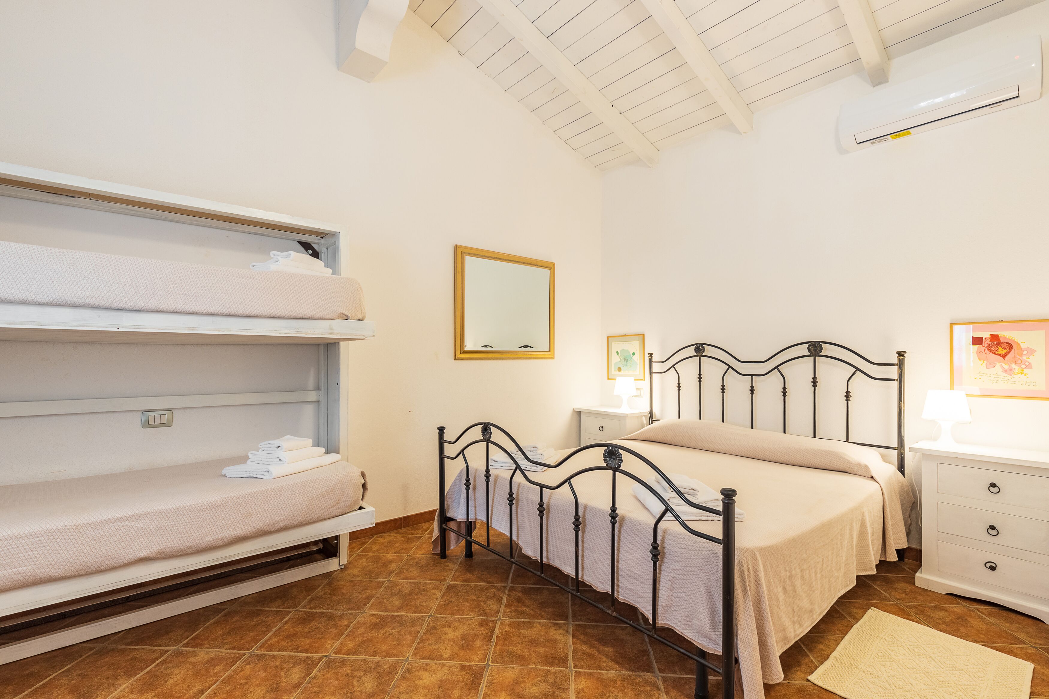 Un dormitorio con una cama litera, una cama individual, una mesita de noche con una lámpara, un mueble auxiliar y un aire acondicionado instalado en la pared.