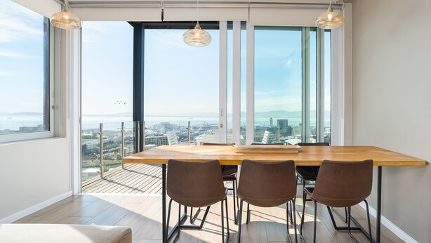 A modern dining area with a large table and four chairs, large windows with a city view, and pendant lights. - High Level 1 (Cape Town)