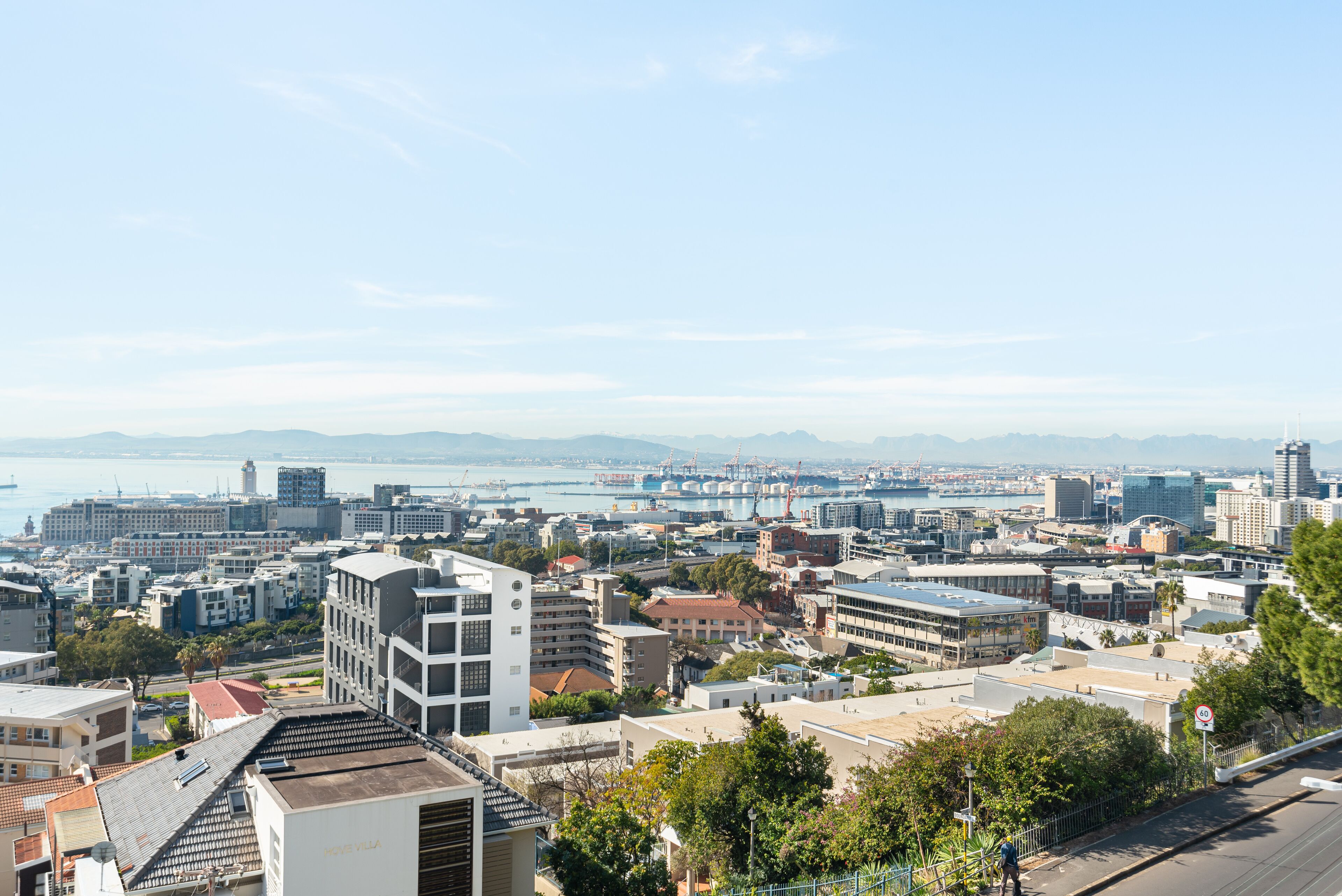 Un paysage urbain où se côtoient des bâtiments modernes et traditionnels, avec une vue dégagée sur la mer et une chaîne de montagnes à l’horizon.