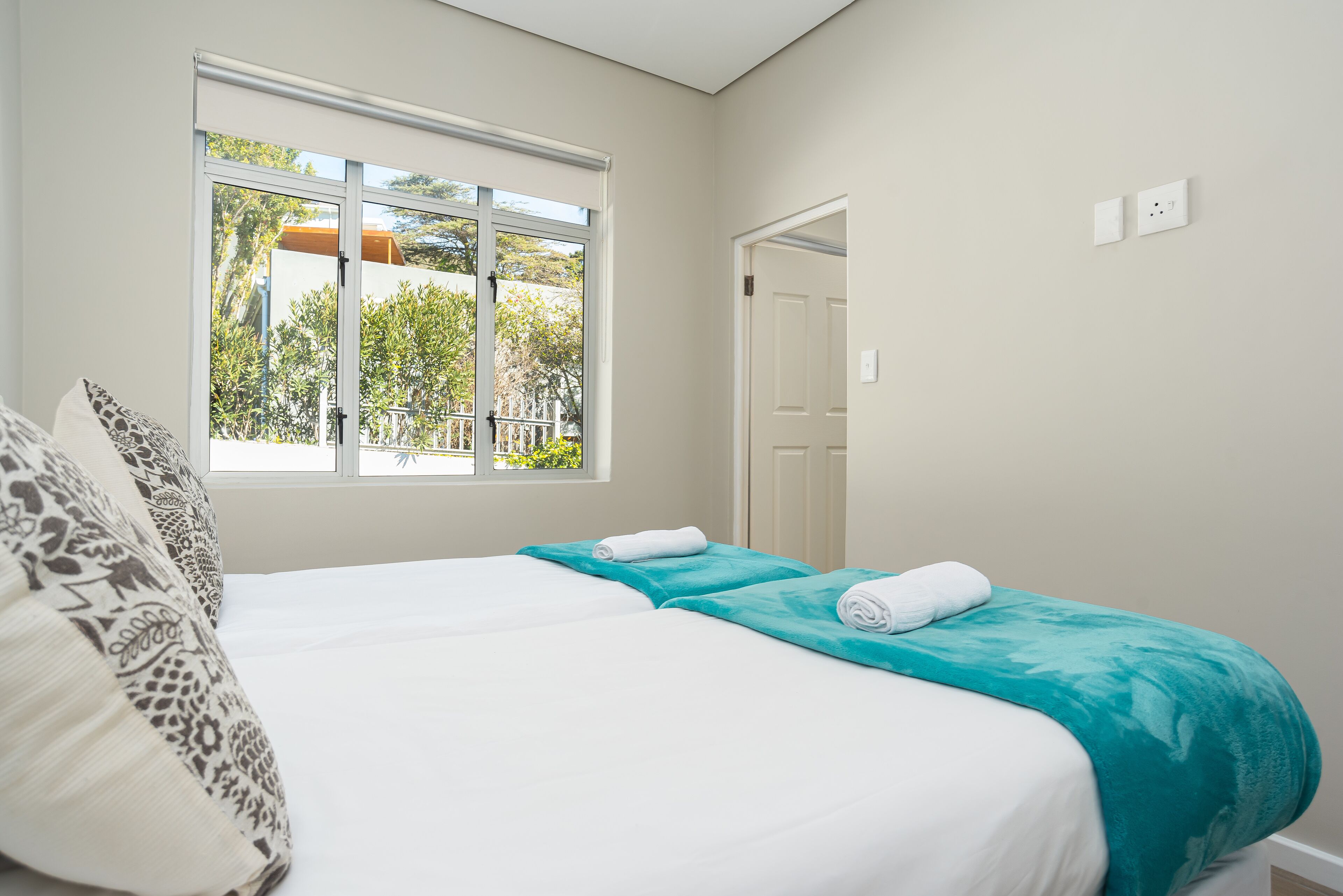 A bedroom with a large bed, a window with a view of greenery, and a door leading to another room.