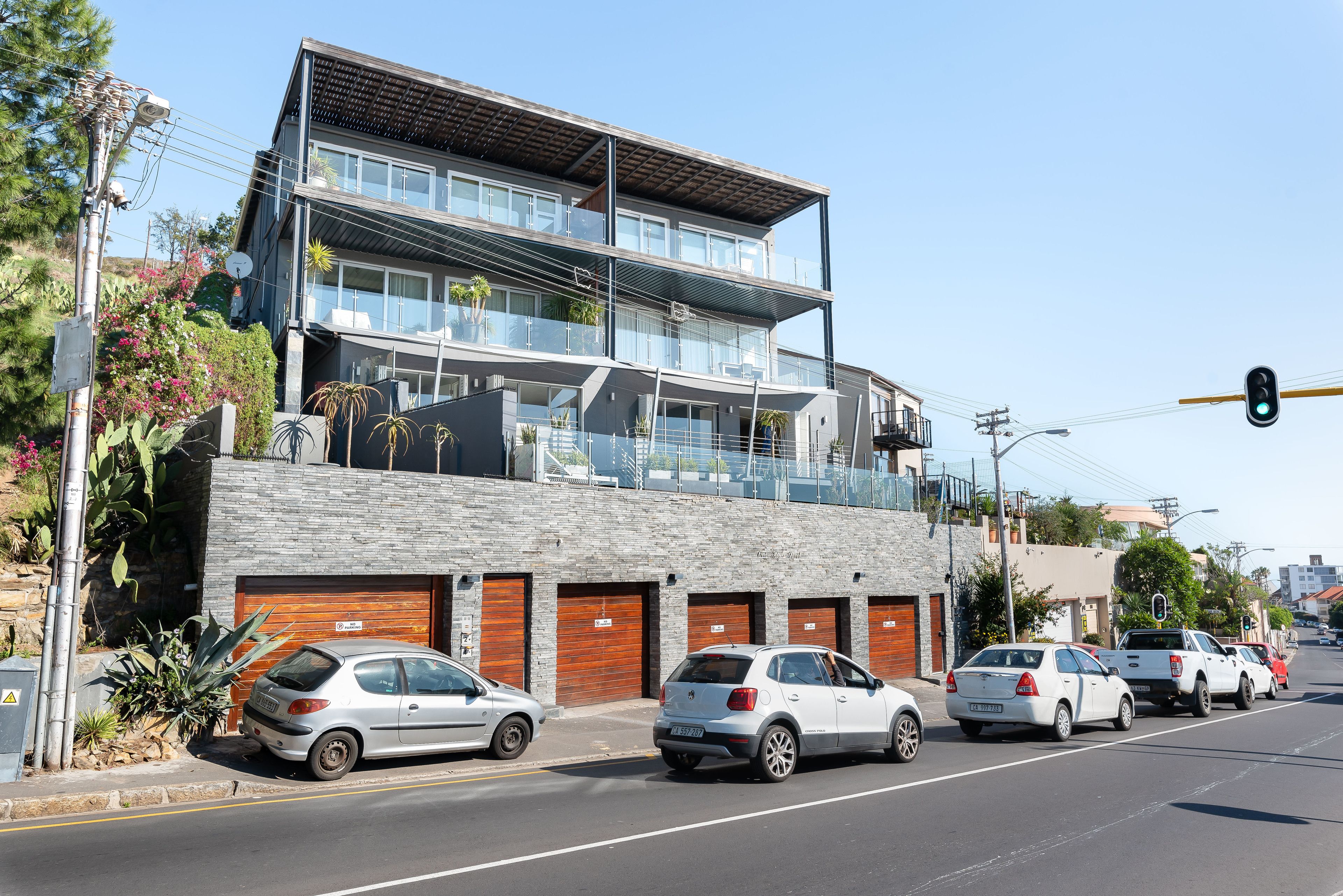 A modern multi-story building with a flat roof, large windows, and balconies. There are several cars parked in front, and a traffic light is visible.
