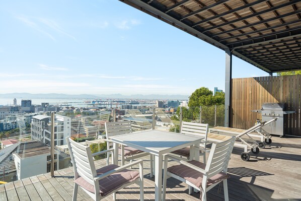 A rooftop terrace with a table and chairs, a grill, and a view of a cityscape.