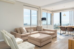 Ett modernt vardagsrum med en beige hörnsoffa, en vit fåtölj och en matplats med ett bord och stolar. Stora fönster ger utsikt över staden och havet.