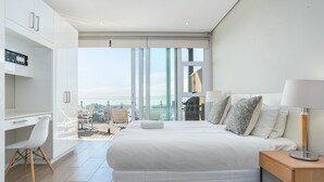 A modern hotel room with a large bed, a desk, and a chair. There is a view of the city through large windows. - High Level 1 (Cape Town)