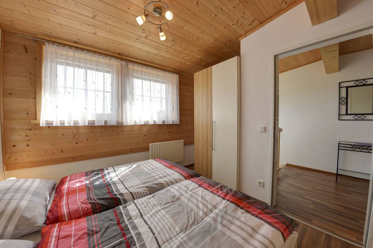Een slaapkamer met een houten plafond, een bed met ruitdessin beddengoed, een raam met dunne gordijnen en een spiegel aan de muur.