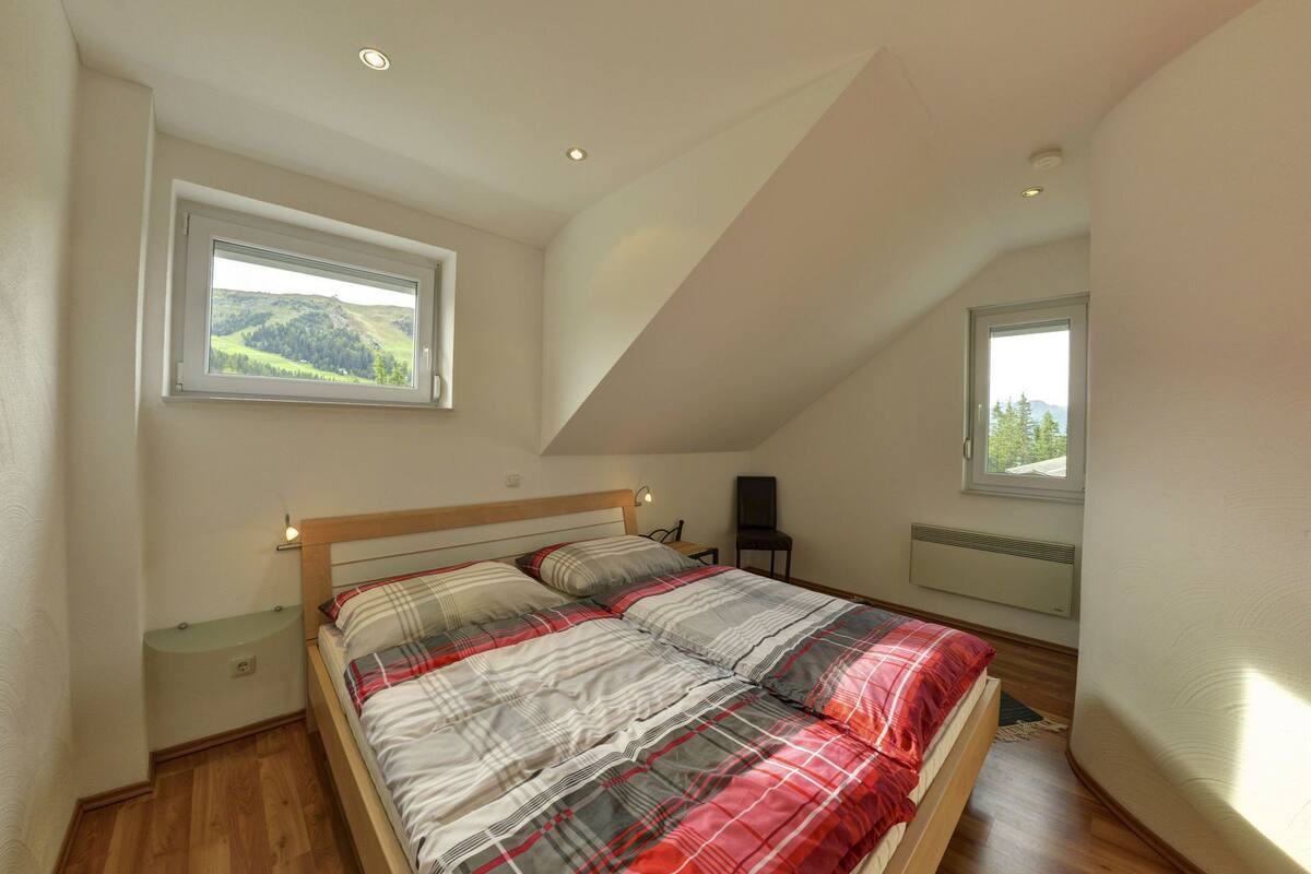 Een slaapkamer met een schuine kap, een bed met ruitjes beddengoed, een stoel, een radiator en een raam met uitzicht op groen.