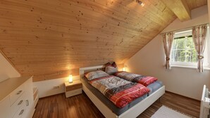 Un dormitorio con techo inclinado, suelo de madera, una cama con ropa de cama estampada, mesitas de noche con lámparas y una ventana con cortinas.