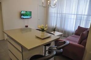 A modern living room with a beige sofa, a dining table with chairs, a TV mounted on the wall, and a window with sheer curtains.