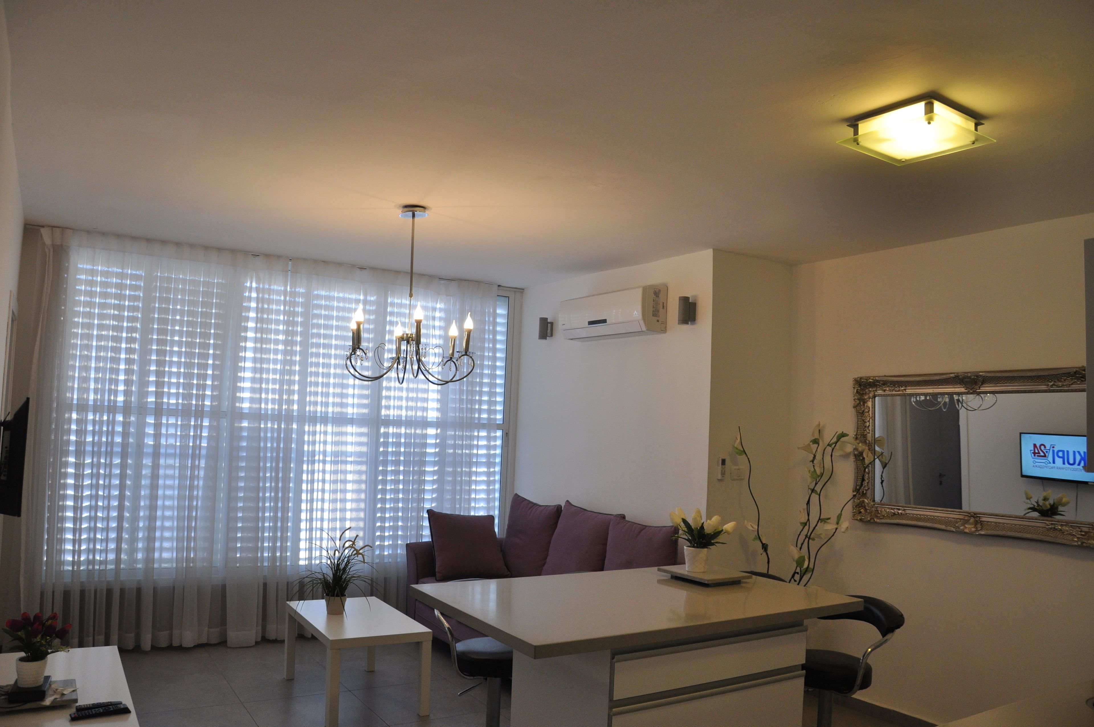 A modern living room with a dining area, a sofa, a chandelier, and a large window with sheer curtains.