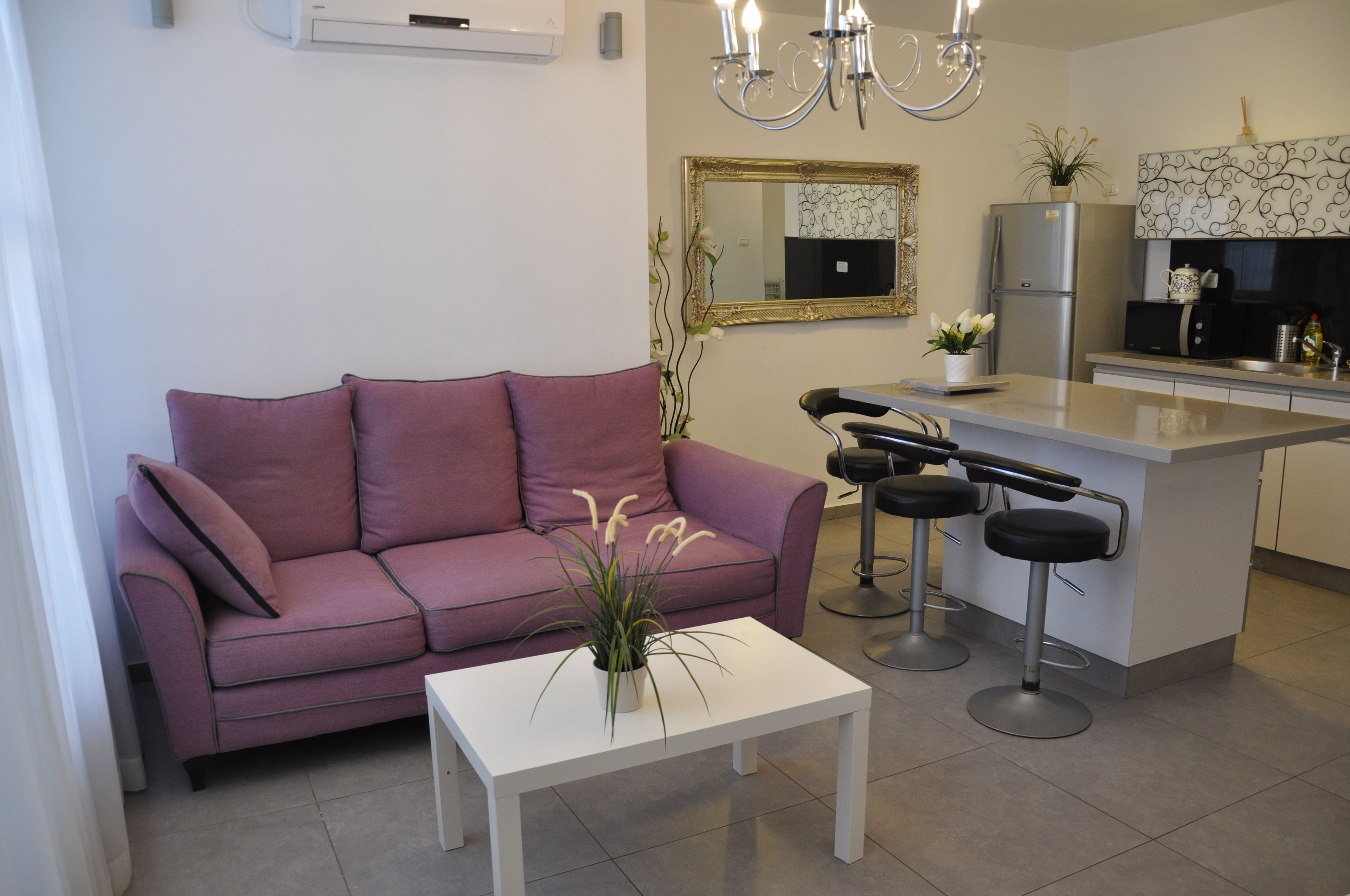 A modern living room with a purple sofa, a white coffee table with a plant, and a kitchen area with a refrigerator, microwave, and bar stools.