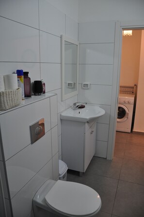 A compact bathroom with a white toilet, a sink, and a mirror. There is a washing machine visible in the adjacent room. - Isra Home Rothschild 8-3 Apartment (Bat Yam)