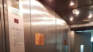 An elevator interior with metallic walls, a control panel with buttons, and a small sign on the wall. - Isra Home Apartment Jabotinsky 8 (Bat Yam)
