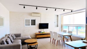 A modern living room with a sofa, coffee table, dining table, and chairs. There is a flat-screen TV mounted on the wall, a wooden cabinet, and a window providing a view of the outdoors. - Isra Home Apartment Jabotinsky 8 (Bat Yam)