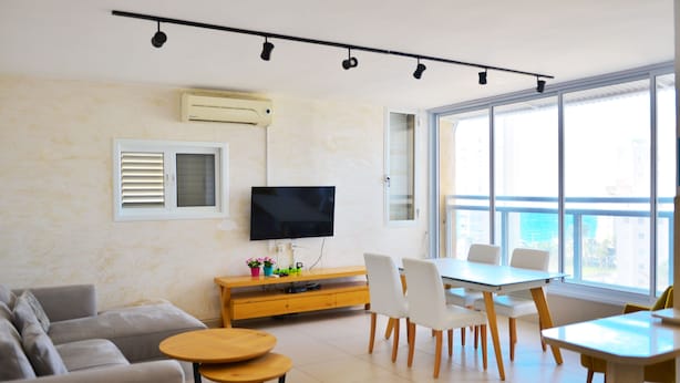 A modern living room with a sofa, a TV, a dining table with chairs, and a large window offering a city view. - Isra Home Apartment Jabotinsky 8 (Bat Yam)