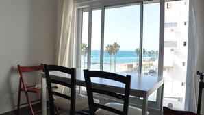 A dining area with a table and chairs, a window with a view of the beach and ocean, and a balcony with a railing. - Isra Home Apartments Ben Gurion 81 (Bat Yam)