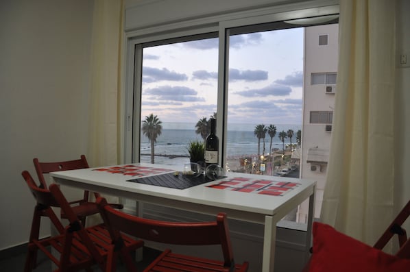 A dining area with a table and chairs, a view of the beach and ocean through large windows, and a balcony with a railing.
