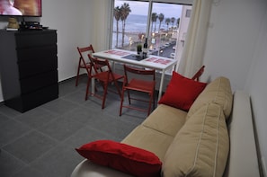 A living room with a beige sofa, red cushions, a dining table with chairs, a TV on a black cabinet, and a view of the beach and palm trees through a window.