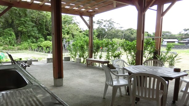 Un patio techado al aire libre con una mesa y sillas, rodeado de follaje y vistas del paisaje.