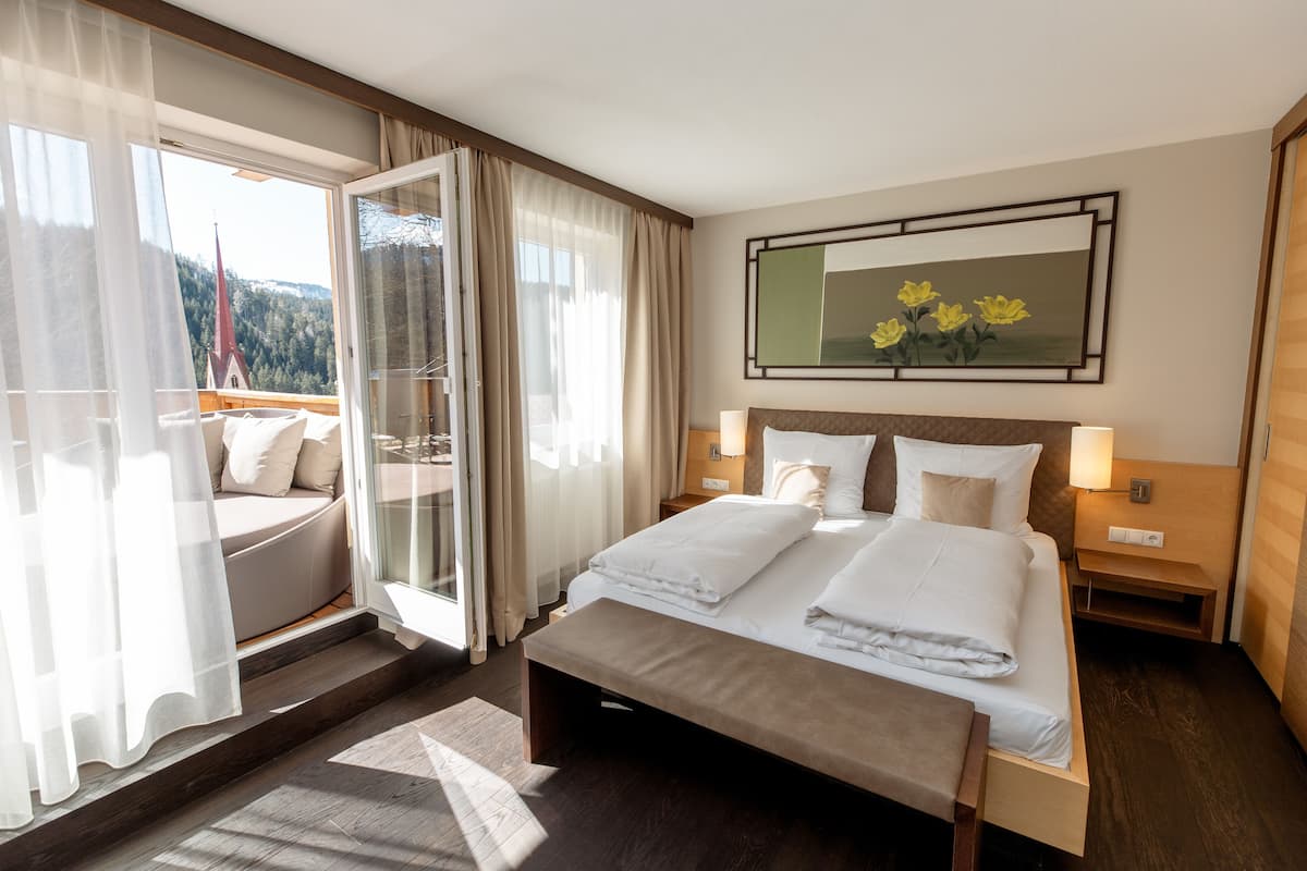 A hotel room with a large bed, a bench, a glass door leading to a balcony, and a painting on the wall.