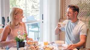 A dining table with breakfast items, a window with curtains, and a view of greenery outside. - Hotel Marica (Nova Ponente)
