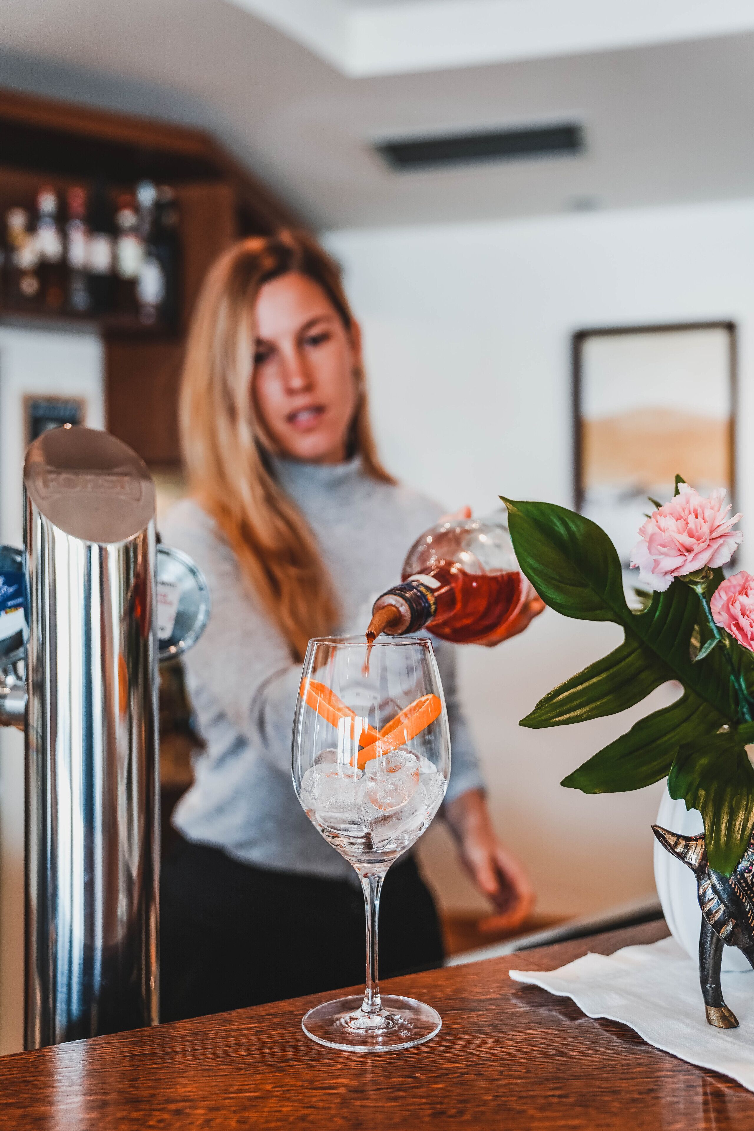 A person pouring a drink into a glass with ice cubes, a cocktail shaker, and a vase with flowers on a wooden table.
