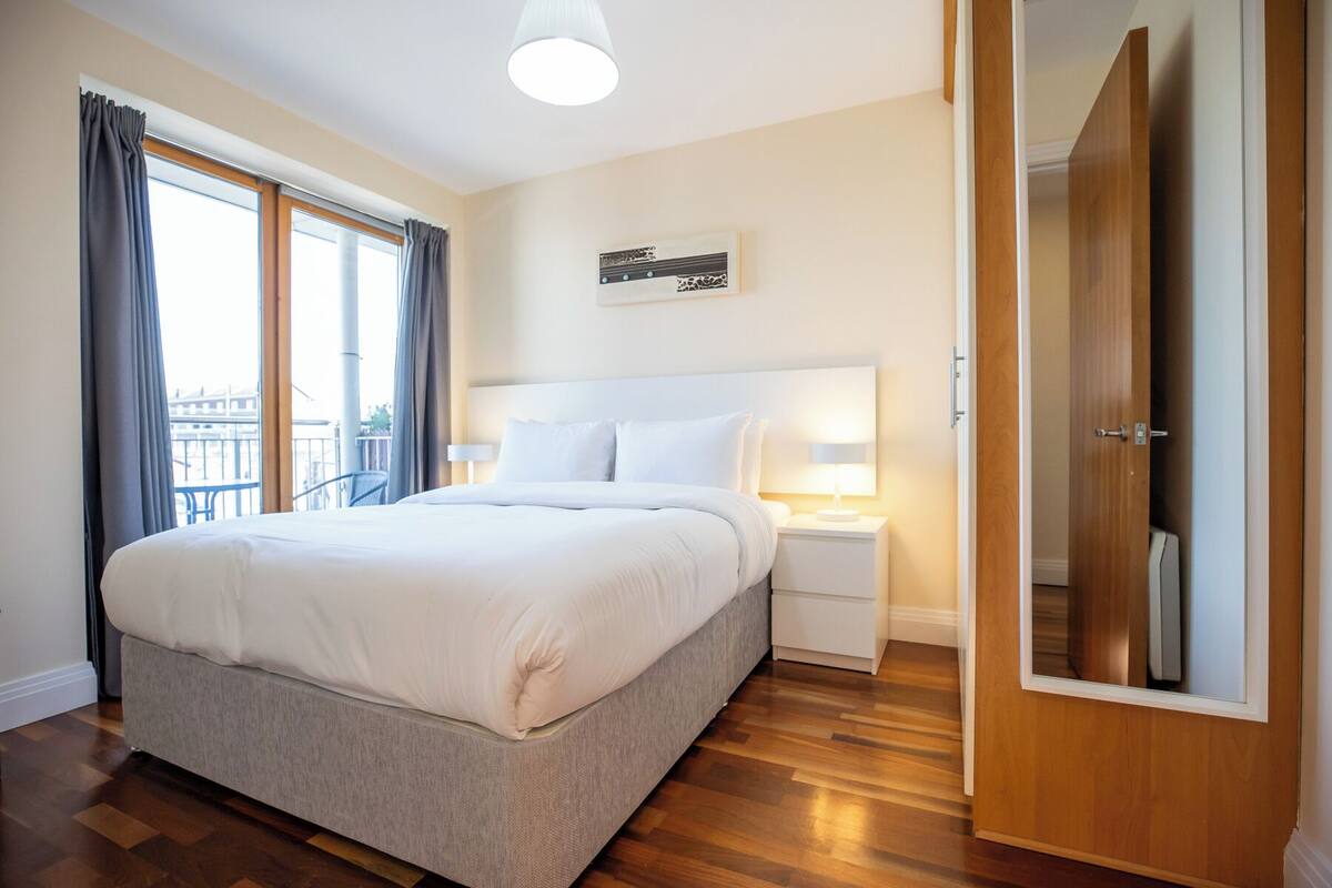 Habitación de hotel con una cama grande, una mesita de noche, una lámpara, un armario y una ventana con cortinas.