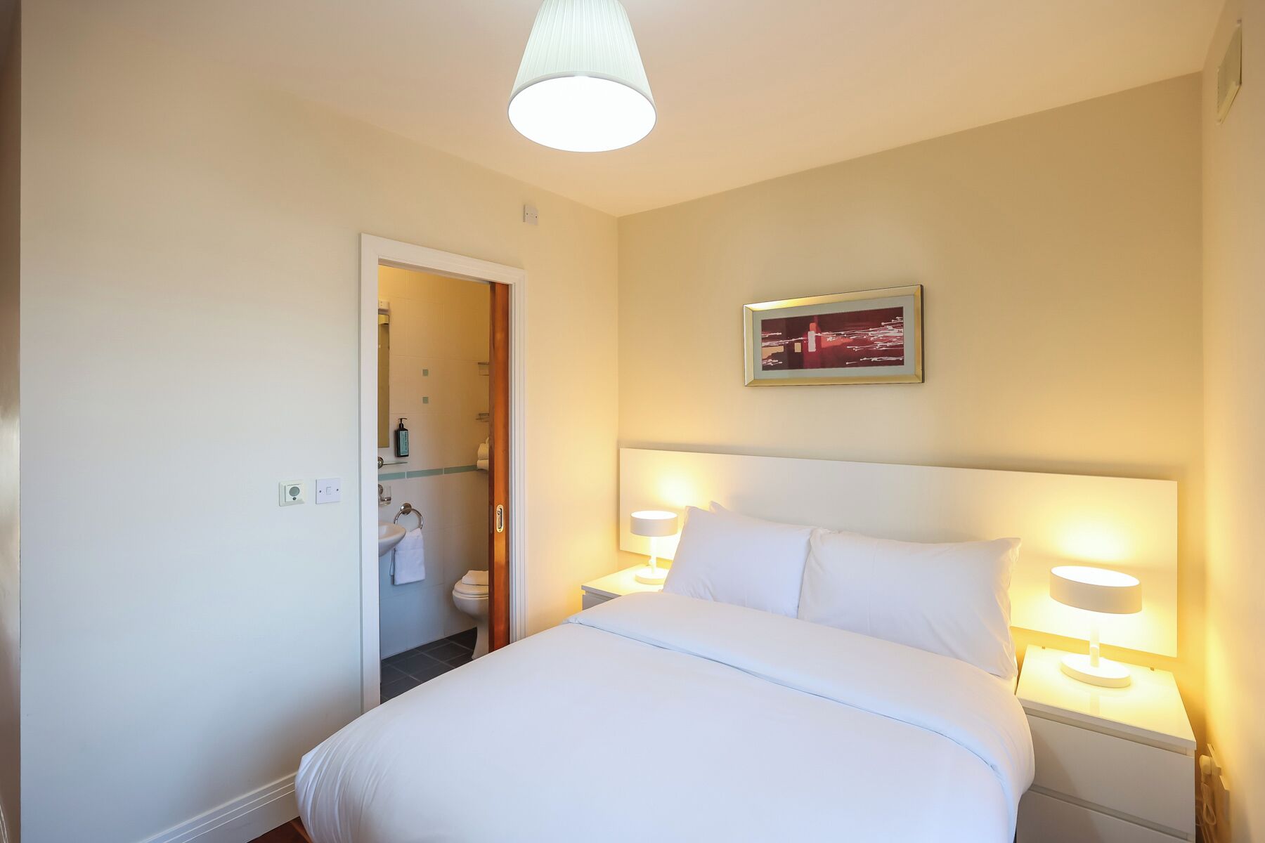 A hotel room with a bed, bedside lamps, a nightstand, a framed picture on the wall, and a bathroom visible through an open door.