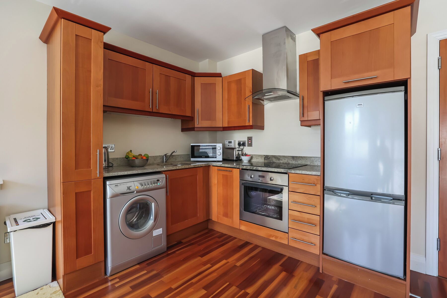 A compact kitchen with wooden cabinets, a washing machine, a microwave, an oven, and a refrigerator.