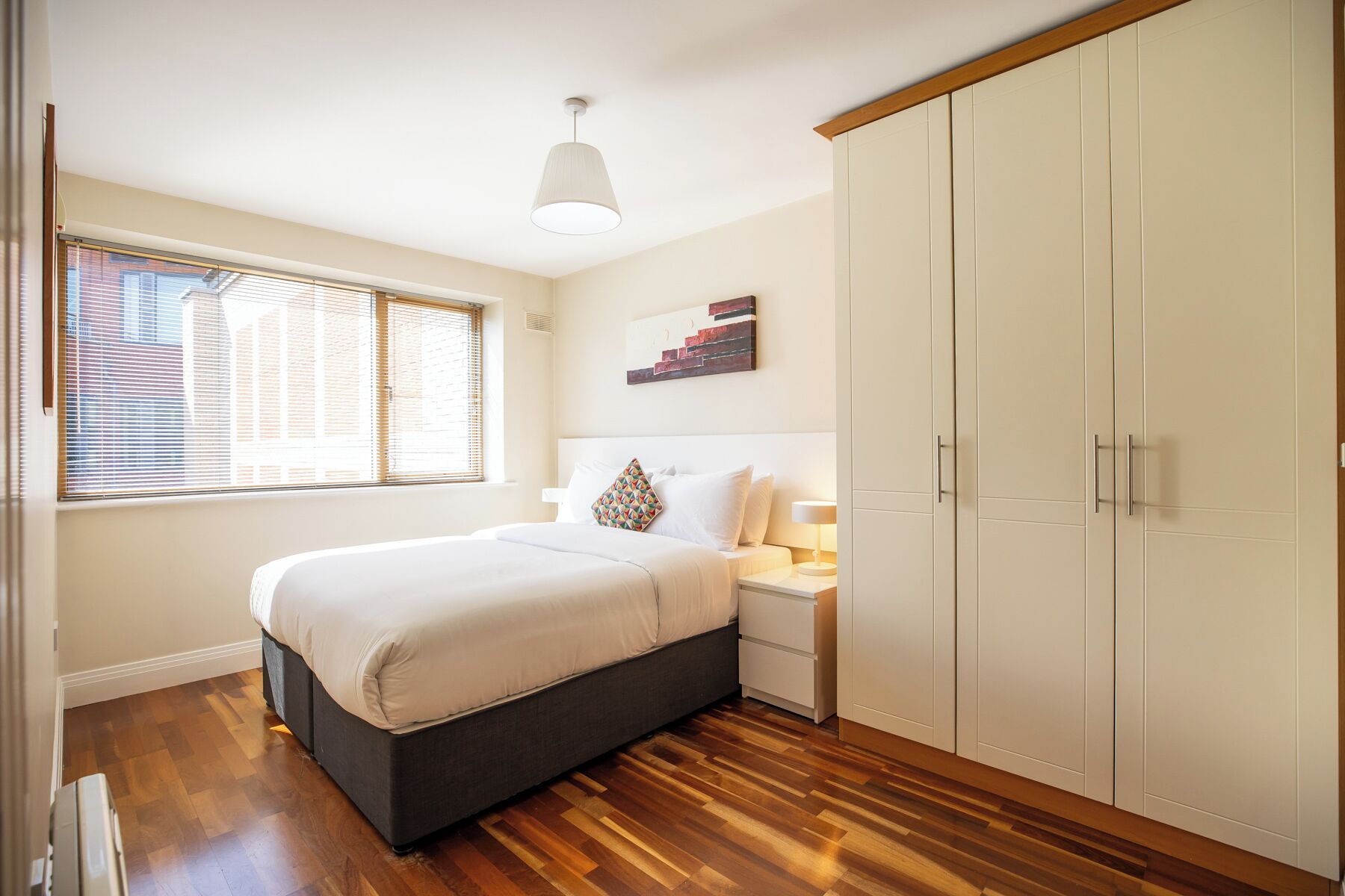 A bedroom with a bed, bedside table, lamp, wardrobe, and window with blinds.