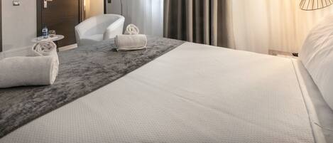 Habitación de hotel con una cama grande, dos sillas blancas, una mesita redonda con una botella de agua y un vaso, y una toalla sobre la cama.