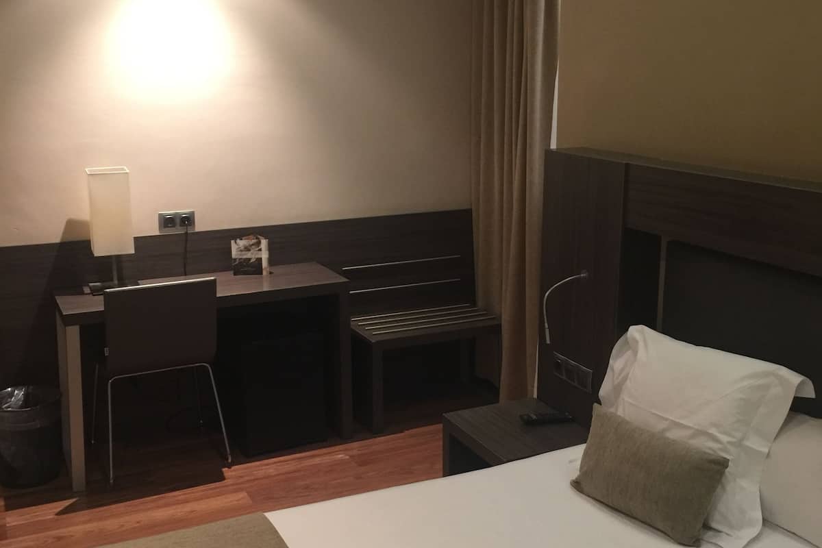 Hotellihuone, jossa on sänky, työpöytä, tuoli, lamppu ja ikkuna, jossa on verhot.