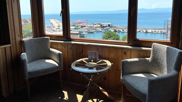 A cozy room with two upholstered chairs and a small glass table, overlooking a marina with boats and a distant coastline. - Denizci Hotel (Sinop)