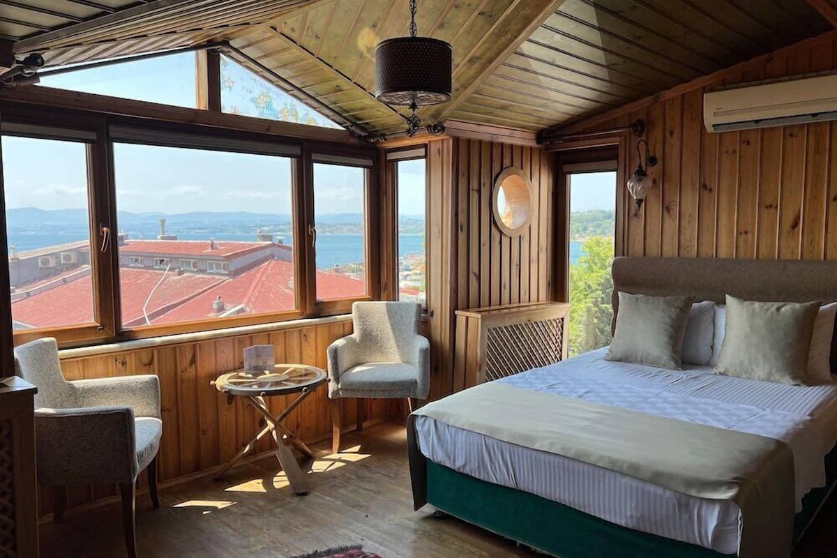 A bedroom with a large bed, a chair, a small table, and a view of the city through large windows.