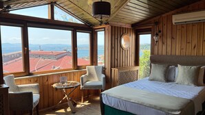 A wooden room with a bed, two chairs, a small table, and large windows offering a view of buildings and water. - Denizci Hotel (Sinop)